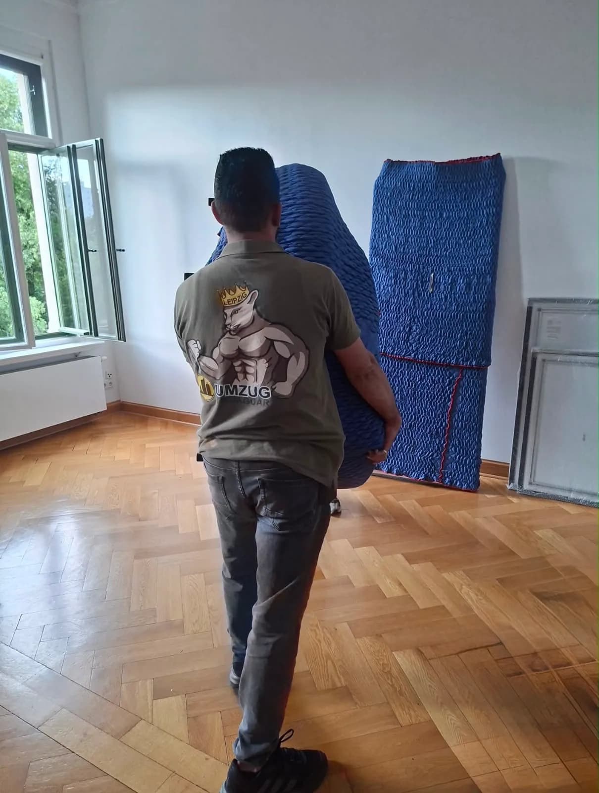 Umzugshelfer trägt eine verpackte Matratze durch einen leeren Altbauraum