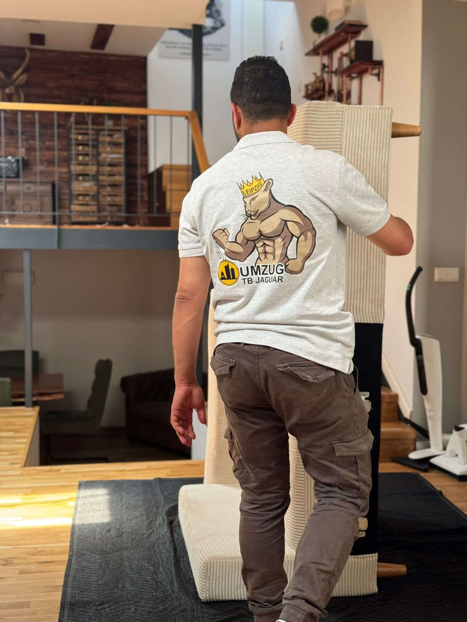 Ein Mann in einem hellgrauen "Umzug TB Jaguar"-Shirt mit Jaguar-Logo trägt einen Teppich oder eine Matte die Holztreppe in einer modernen, offenen Wohnung hinauf und demonstriert damit die Professionalität der Dienstleistungen des Firmenumzugs Leipzig. - Umzug TB Jaguar Leipzig